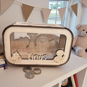 May include: A wooden money box with a clear front panel. The box features laser-cut animal illustrations, including an elephant, giraffe, lion, and monkey. The name "Linus" is also cut out. Several coins are visible in front of the box.