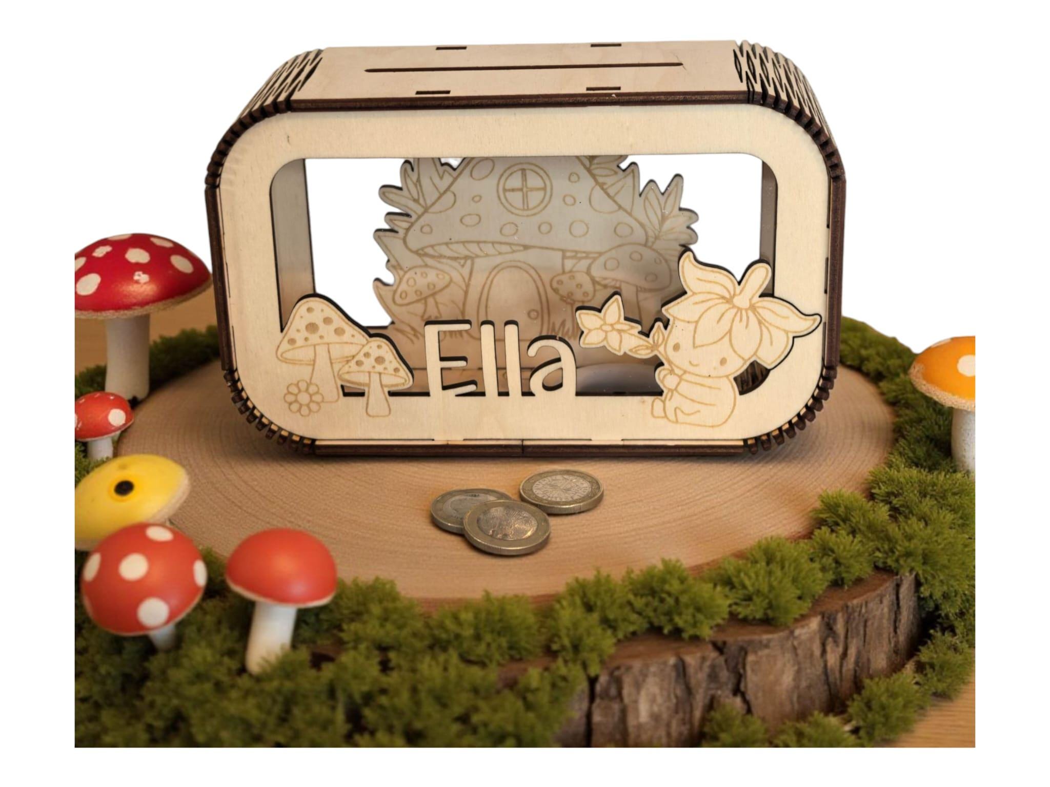 May include: A wooden money box with a fairy-tale theme. The box features a laser-cut design of a mushroom house, mushrooms, and a flower fairy. The name "Ella" is also laser-cut on the front. The box is surrounded by decorative mushrooms and moss.