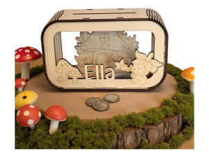 May include: A wooden money box with a fairy-tale theme. The box features a laser-cut design of a mushroom house, mushrooms, and a flower fairy. The name "Ella" is also laser-cut on the front. The box is surrounded by decorative mushrooms and moss.